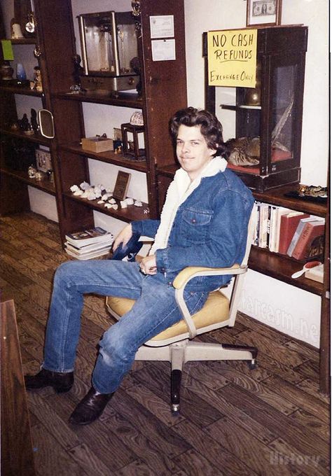 A man in a blue jacket with blue jeans sitting on a brown desk chair in front of a wall with brown shelves
